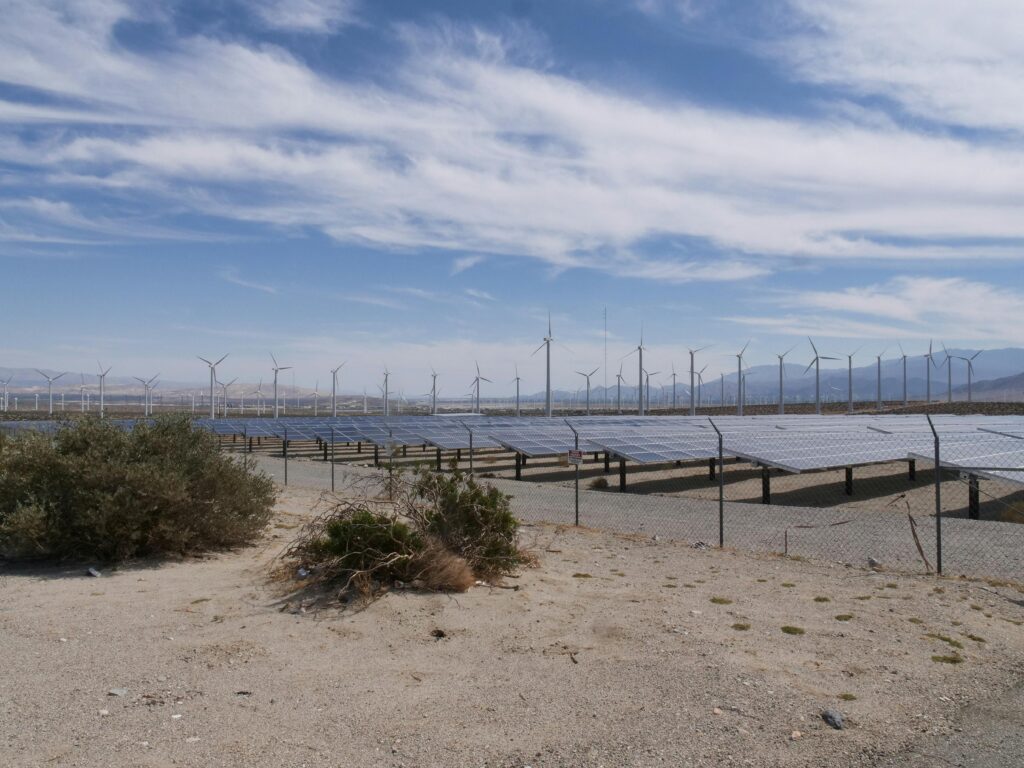 Expansive solar panels and wind turbines in a desert landscape under a bright blue sky.